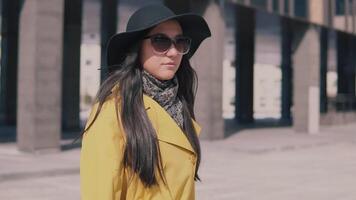 elegante ragazza nel un' giallo cappotto con lungo buio capelli nel un' cappello passeggiate giù il strada prende via il suo bicchieri e sembra intorno a. lento movimento video