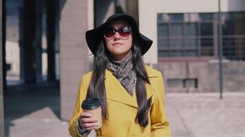 elegante niña en un amarillo impermeable sombrero y lentes camina abajo el calle y bebidas café. lento movimiento video