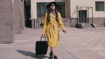 stylish young girl with long dark hair in a yellow raincoat hat and glasses comes with luggage video