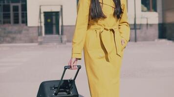 lindo mulher com grandes Sombrio cabelo dentro uma amarelo capa e óculos vem com bagagem. lento movimento video