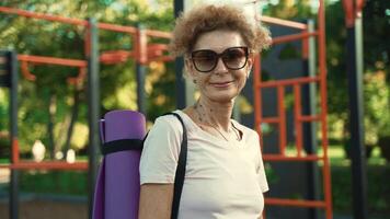 Portrait of senior woman standing in front of outdoor sports ground with yoga mat on shoulder in summer sunshine with smile on face. Fitness and leisure activities in open space. Mature activity. video