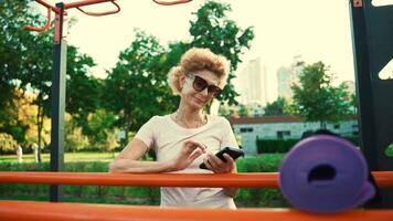 sportivo maturo donna usi Telefono a strada Palestra dopo yoga classe. anziano caucasico signora assunzione rompere dopo yoga pratica all'aperto. attivo stile di vita concetto. le persone, sport e tecnologia. video