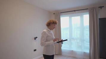 Adult caucasian woman dressed business style works as real estate agent, stands in bright new apartment, looks at design options on tablet while waiting for clients, holds the keys in her hand. video