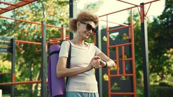 Senior woman with yoga mat stands on street gym and uses sports watch. An elderly woman holding a yoga pad and checking her wristwatch after practice. Modern sports gadgets, fitness equipment concept. video