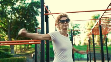 Older female warms up before working out on street fitness site in summer. Elderly woman doing warm-up exercise for arms and upper body belt on outdoor workout area. Retirement healthy way of living. video