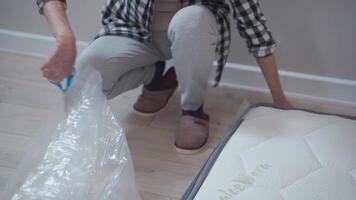 Topic of moving to new apartment. An elderly woman unpacks a new polyethylene orthopedic mattress in an empty apartment. A happy middle-aged female opens the packaging of a new mattress in a new home. video