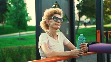 reifen Frau ruhen nach ausüben auf Straße Sport Boden halten Flasche mit Wasser im Sommer. älter weiblich entspannend nach Sport Session auf draussen Spielplatz. aktiv Lebensstil im Erwachsensein. video