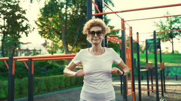 Mature woman warming up before workout at outdoor gym in summer. Elderly female doing exercises to warm up arms and upper shoulder zone at the street fitness area. Healthy lifestyle in retirement. video