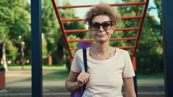 reifen Frau gehen zu Yoga Klasse draußen. aktiv Senior genießen gesund Lebensstil. Senior weiblich halten Übung Matte während Gehen beim Ausbildung Sitzung. positiv Alten Frau mit Sport Ausrüstung. video