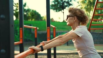 Cheerful active mature lady is training her upper body, doing push-ups from the horizontal bar on the street in the gym. Elderly woman doing arm exercises do pushups outdoors at the fitness area. video