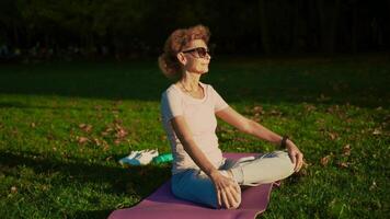 yoga à parc. milieu âge femme dans lotus pose séance sur yoga tapis sur vert herbe. concept de calme et méditation. yoga en plein air. Sénior femme méditer et exercice yoga lotus position dans la nature. video