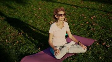 senior kvinna mediterar och utövar yoga lotus placera utomhus. ederly kvinna håller på med stretching övningar på yoga matta i parkera på grön gräsmatta på solnedgång. känsla energi. begrepp lugna och meditation. video