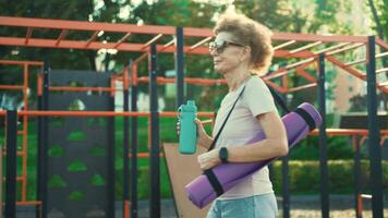 reifen Frau gehen zu Yoga Klasse draußen. aktiv Senior genießen gesund Lebensstil. Senior weiblich halten Übung Matte während Gehen beim Ausbildung Sitzung. positiv Alten Frau mit Sport Ausrüstung. video