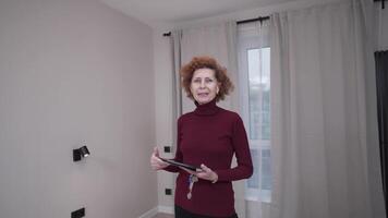 uthyrning och försäljning av hus. en mogna kvinna verklig egendom ombud visar av ny tömma lägenhet medan ser på de kamera. porträtt av en fastighetsmäklare i ett tömma lägenhet. reklam fast egendom innan försäljning. video