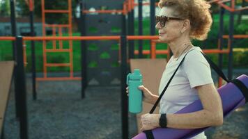 Active senior woman with fitness mat and water bottle in hands walking on workout outdoor gym. Happy mature lady strolling in sports ground going at outdoors yoga class. Active lifestyle concept. video