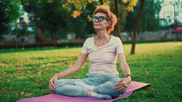 senior vrouw mediteren en oefenen yoga lotus positie buitenshuis. ederly vrouw aan het doen uitrekken opdrachten Aan yoga mat in park Aan groen gazon Bij zonsondergang. gevoel energie. concept kalmte en meditatie. video