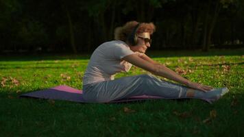 caucasico in forma donna matura fare esercizi per caldo su e allungare muscoli su un' yoga stuoia nel parco, ascoltando per musica nel cuffia. concetto di un' salutare stile di vita su la pensione. sport nel il anziano. video