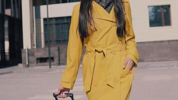 stylish woman in a yellow raincoat with long dark hair goes with luggage removes glasses looks around and smiles. slow motion video