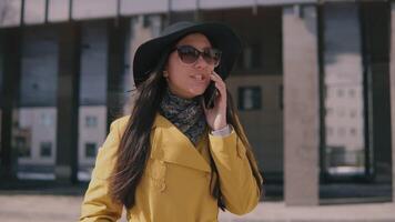 stylish young girl with long dark hair in a yellow raincoat hat and glasses talking on the phone and laughing video