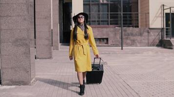 stylish girl in a yellow raincoat hat and glasses goes with luggage and smiles. slow motion video