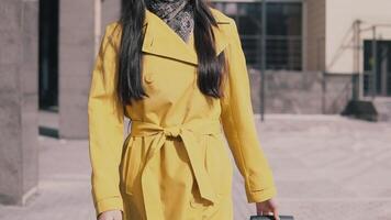 stylish woman in a yellow raincoat hat and glasses goes with luggage and smiles. slow motion video