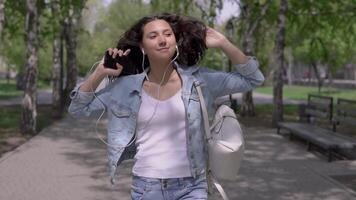 gracioso niña con largo pelo en un mezclilla chaqueta y un mochila va abajo el calle y bailes escuchando a música en auriculares utilizando un teléfono inteligente video