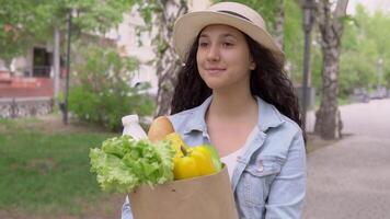 ein jung schön Frau im ein Denim Jacke und ein stilvoll Hut mit ein gut Stimmung Spaziergänge Nieder das Straße im ein Stadt Park und trägt ein groß Paket von frisch Produkte und lächelt. video