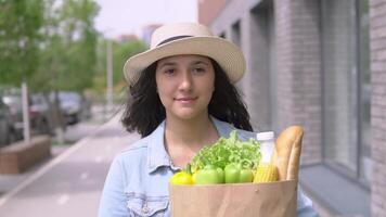 un joven hermosa mujer en un mezclilla chaqueta y sombrero camina abajo el calle y lleva un bolso de Fresco sabroso productos el concepto de apropiado nutrición. compras, sano comida productos video