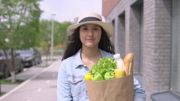 een jong mooi vrouw in een denim jasje en hoed wandelingen naar beneden de straat in een mooi zo humeur en draagt een zak van vers smakelijk producten. de concept van gepast voeding. winkelen, gezond voedsel producten. video