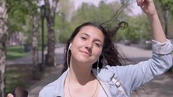 divertente ragazza con lungo capelli nel un' denim giacca e un' zaino a piedi giù il strada e danza mentre ascoltando per musica nel cuffie utilizzando un' smartphone. avvicinamento video