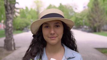 un joven hermosa mujer en un mezclilla chaqueta y un elegante sombrero con un bueno estado animico camina abajo el calle en un ciudad parque y lleva un grande paquete de Fresco productos y sonrisas video