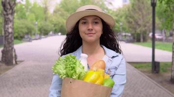 A young attractive woman in a denim jacket and hat walks down the street in a good mood and carries a bag of fresh tasty products. video