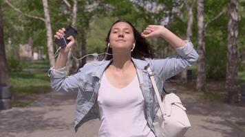 engraçado menina com grandes cabelo dentro uma jeans Jaqueta e uma mochila anda em baixa a rua e danças engraçado ouvindo para música dentro fones de ouvido usando uma smartphone.lento movimento video