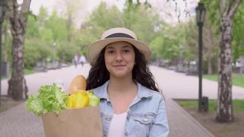 een jong mooi vrouw in een denim jasje en elegant hoed wandelingen naar beneden de straat in een stad park en draagt een groot pakket van producten. video