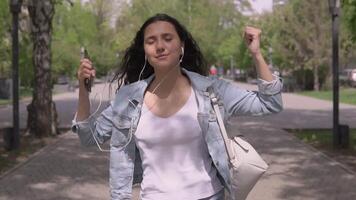 funny girl with long hair in a denim jacket and a backpack goes down the street and dances listening to music in headphones using a smartphone.slow motion video