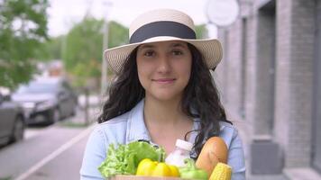 A young attractive woman in a denim jacket and hat walks down the street in a good mood and carries a bag of fresh tasty products. video