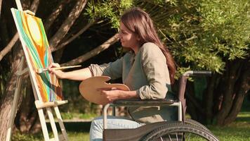 une désactivée femme dans une fauteuil roulant des peintures une image dans le parc dans ensoleillé été temps. désactivée personne, femme artiste, art pour ventes, inspiration dans la nature. video