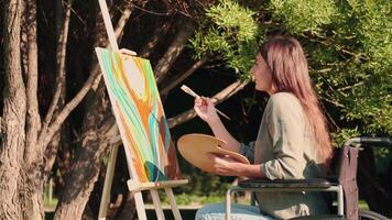 un discapacitado mujer en un silla de ruedas sorteos un imagen en el parque. discapacitado persona, mujer artista, Arte para ventas, inspiración en naturaleza. video