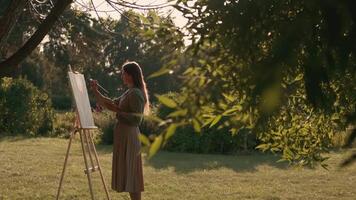 creativo mujer artista pinturas un imagen con un cepillo mientras en pie cerca el caballete en naturaleza. mujer artista, Arte para ventas, inspiración en naturaleza. video