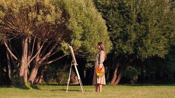 alegre mujer artista pinturas un imagen en lona mientras en pie en un parque en soleado clima. mujer artista, Arte para ventas, inspiración en naturaleza. video