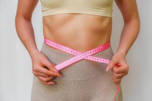 Cropped view of slim woman measuring waist with tape measure at home, close up. An unrecognizable European woman checks the result of a weight loss diet or liposuction indoors. Healthy lifestyle. photo