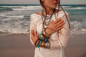 Model in boho style in a white long dress and silver jewelry on the beach. Her hair is braided, and there are many bracelets on her arms. photo