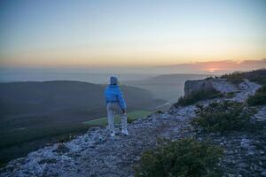 Woman tourist on top of sunrise mountain. The girl salutes the sun, wearing a blue jacket, white hat and white jeans. Conceptual design. photo