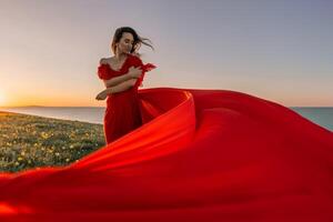 woman red dress standing grassy hillside. The sun is setting in the background, casting a warm glow over the scene. The woman is enjoying the beautiful view and the peaceful atmosphere. photo