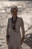 Middle aged woman looks good with blond hair, boho style in white long dress on the beach decorations on her neck and arms. photo