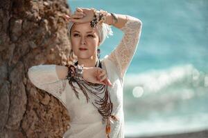 Middle aged woman looks good with blond hair, boho style in white long dress on the beach decorations on her neck and arms. photo