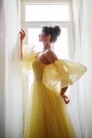 un mujer silueta en un dorado lujoso vestir en contra el antecedentes de un ventana sostiene un cortina con su manos. elegante dama en un amarillo largo seda vestir con desnudo atrás, espalda vista. foto