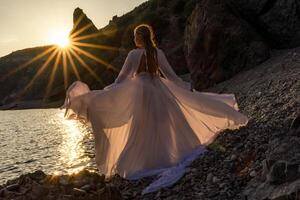 Mysterious woman silhouette long hair walks on the beach ocean water, sea nymph wind listens to the wave. Throws up a long white dress, a divine sunset. Artistic photo from the back without a face