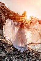 misterioso mujer silueta largo pelo camina en el playa Oceano agua, mar ninfa viento escucha a el ola. lanza arriba un largo blanco vestido, un adivinar puesta de sol. artístico foto desde el espalda sin un cara