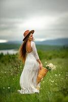 A middle-aged woman in a white dress and brown hat stands on a green field and holds a basket in her hands with a large bouquet of daisies. In the background there are mountains and a lake. photo